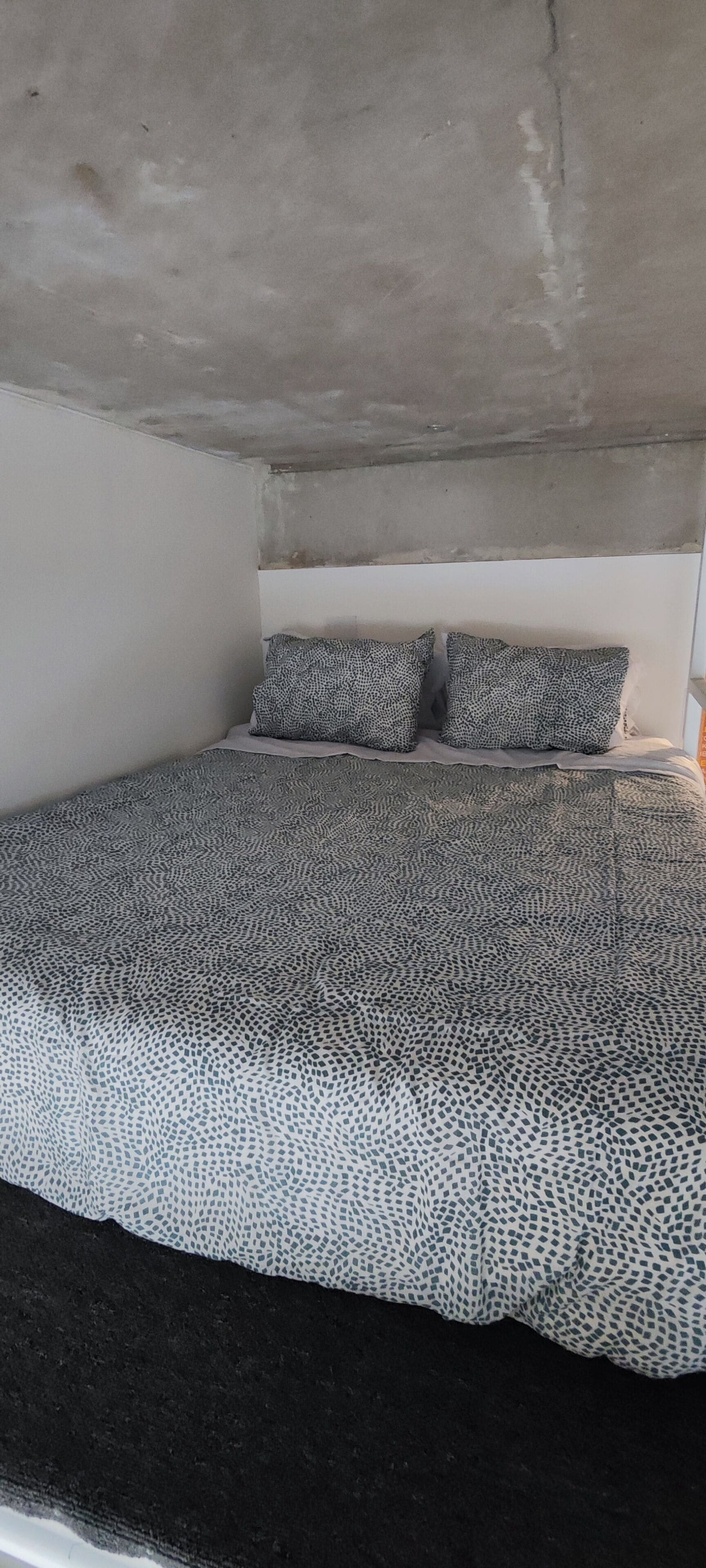 Una habitación pequeña con cama individual, colcha estampada, dos almohadas y techo de hormigón.