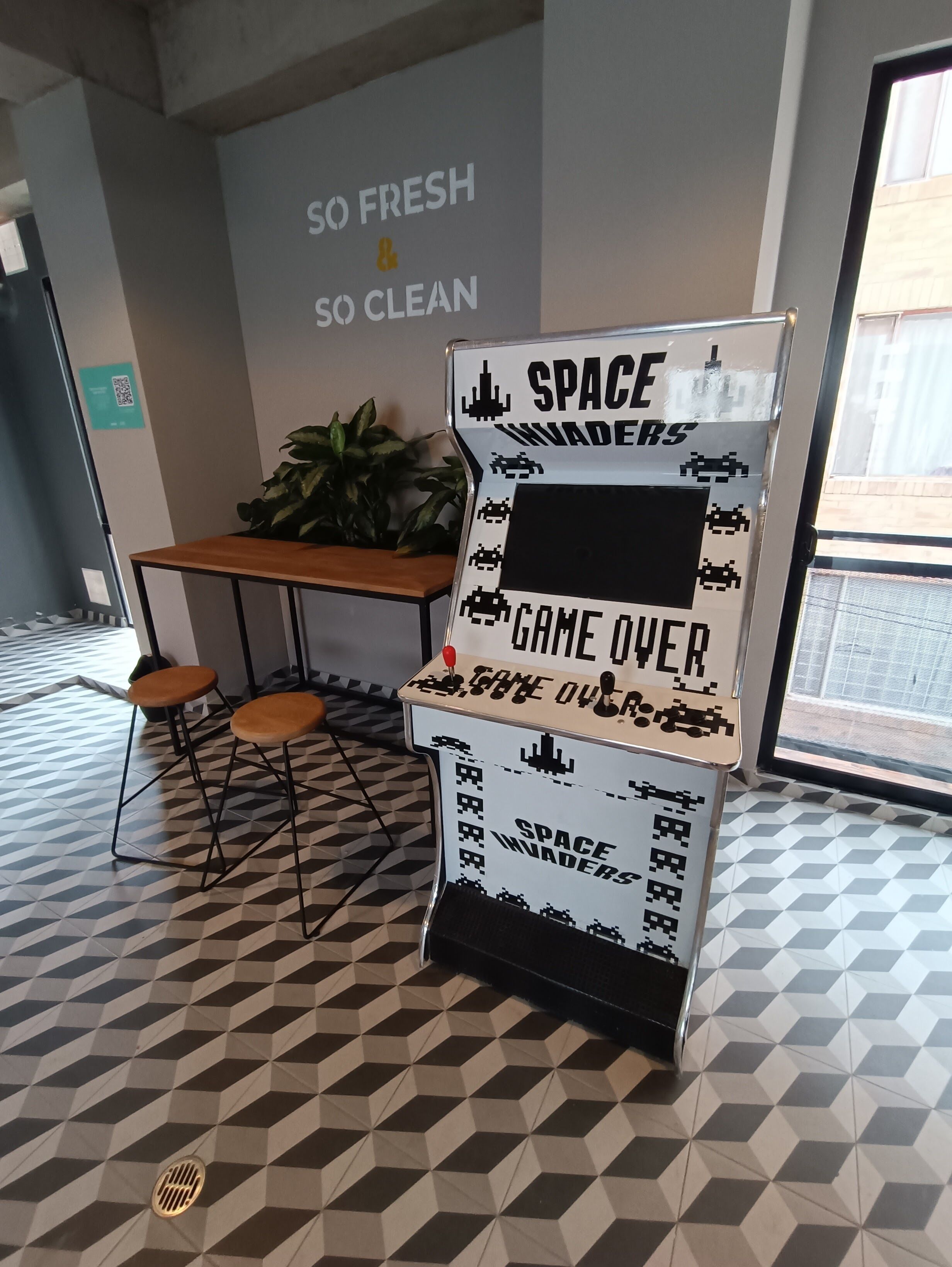A space-themed game station with a sign reading "SPACE INVADERS GAME OVER" in a room with geometric patterned flooring and a wooden table with stools.