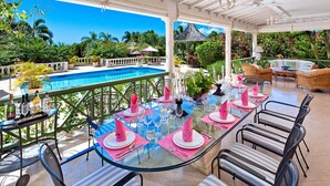 Un espace extérieur abrité, aménagé pour les repas, avec une table en verre dressée, entouré de meubles en osier et offrant une vue sur une piscine et une végétation luxuriante.