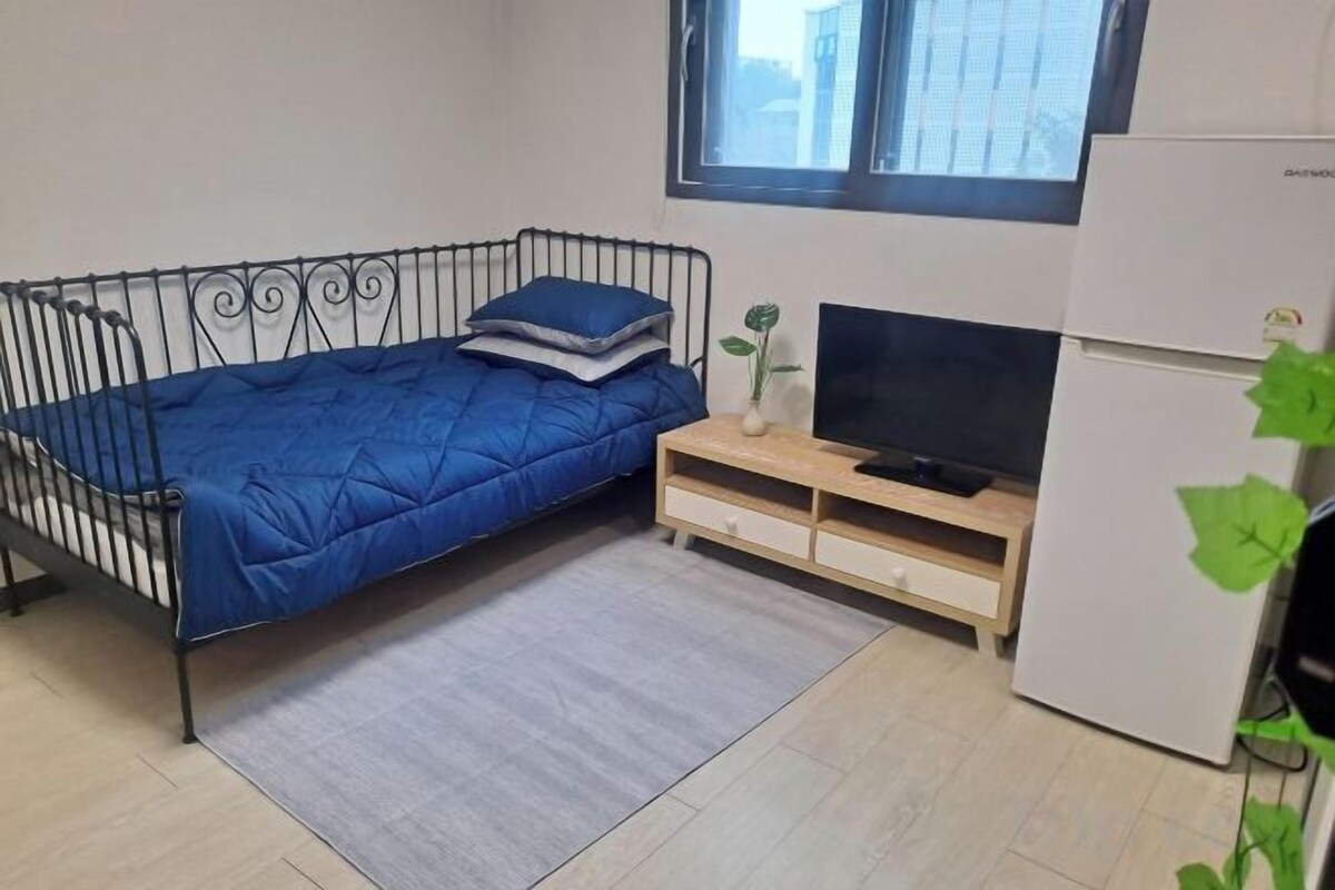 Een kleine kamer met een bed met metalen frame, een televisie op een houten standaard, een koelkast en een raam.