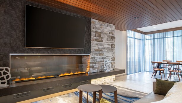 A modern living room with a large flat-screen TV, a stone fireplace, and a dining area with wooden furniture. - Fairfield Inn & Suites Indianapolis Plainfield (Plainfield)