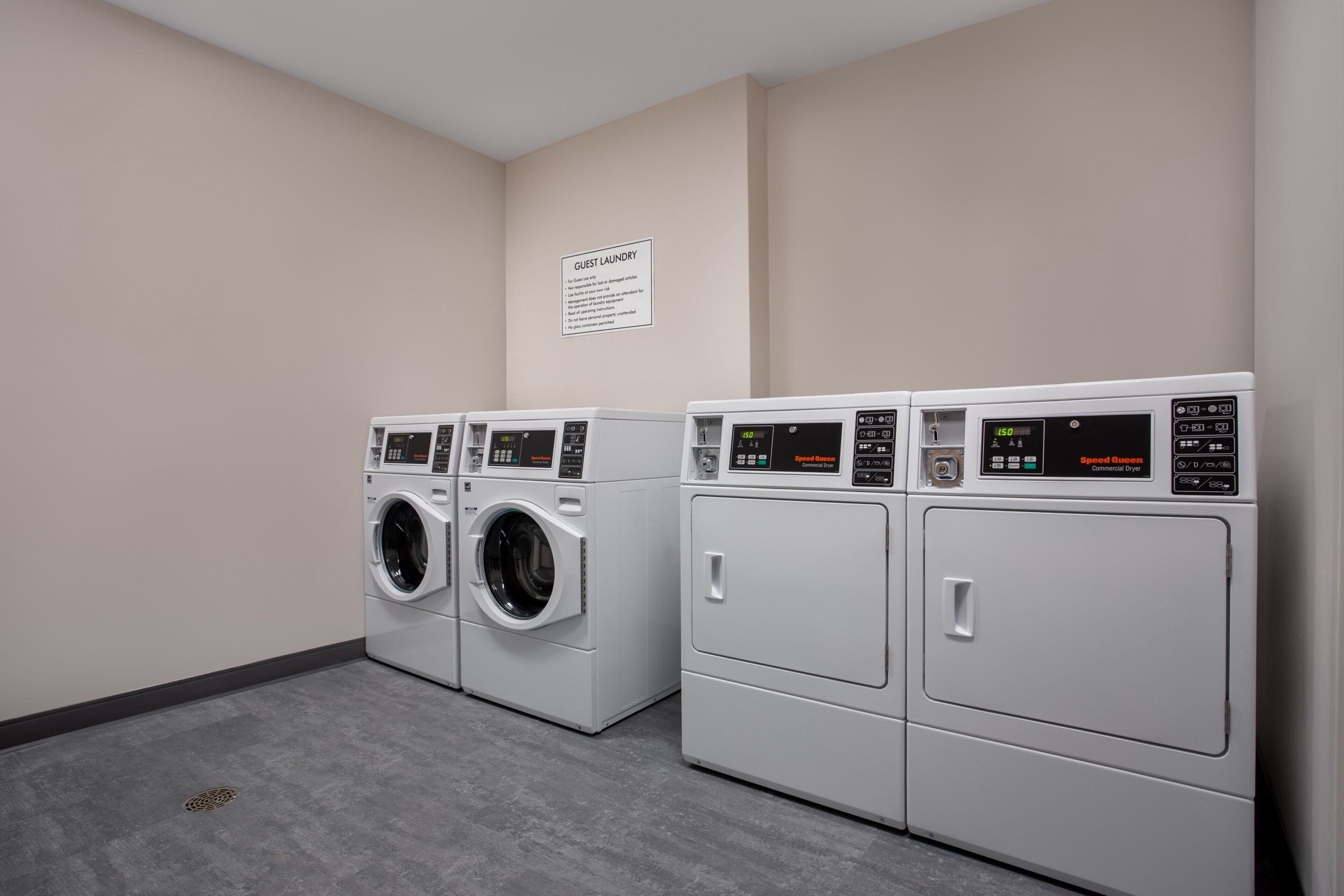 A laundry area with three washers and dryers, a sign indicating guest laundry services, and a grey floor.