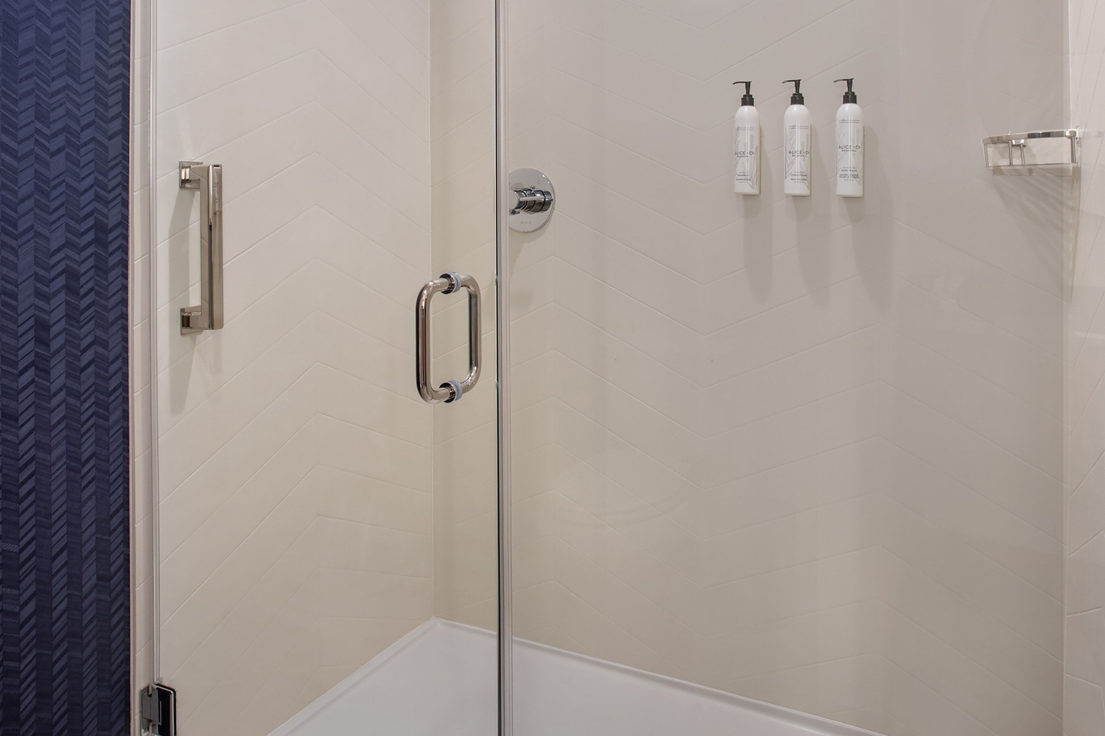 A glass-enclosed shower area with a white tiled wall, a silver towel rack, and three bottles of shampoo on the wall.