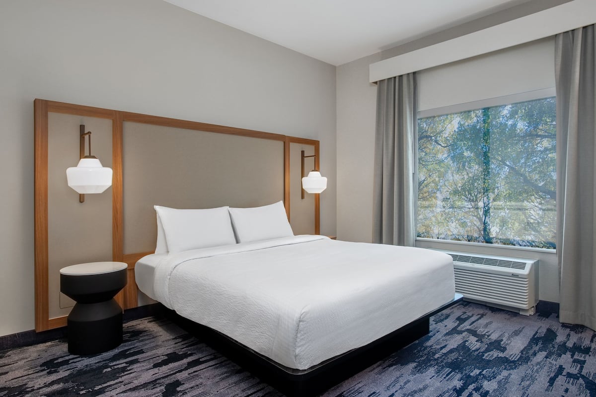 A hotel room with a large bed, a bedside table, a wall-mounted lamp, a window with curtains, and a radiator.
