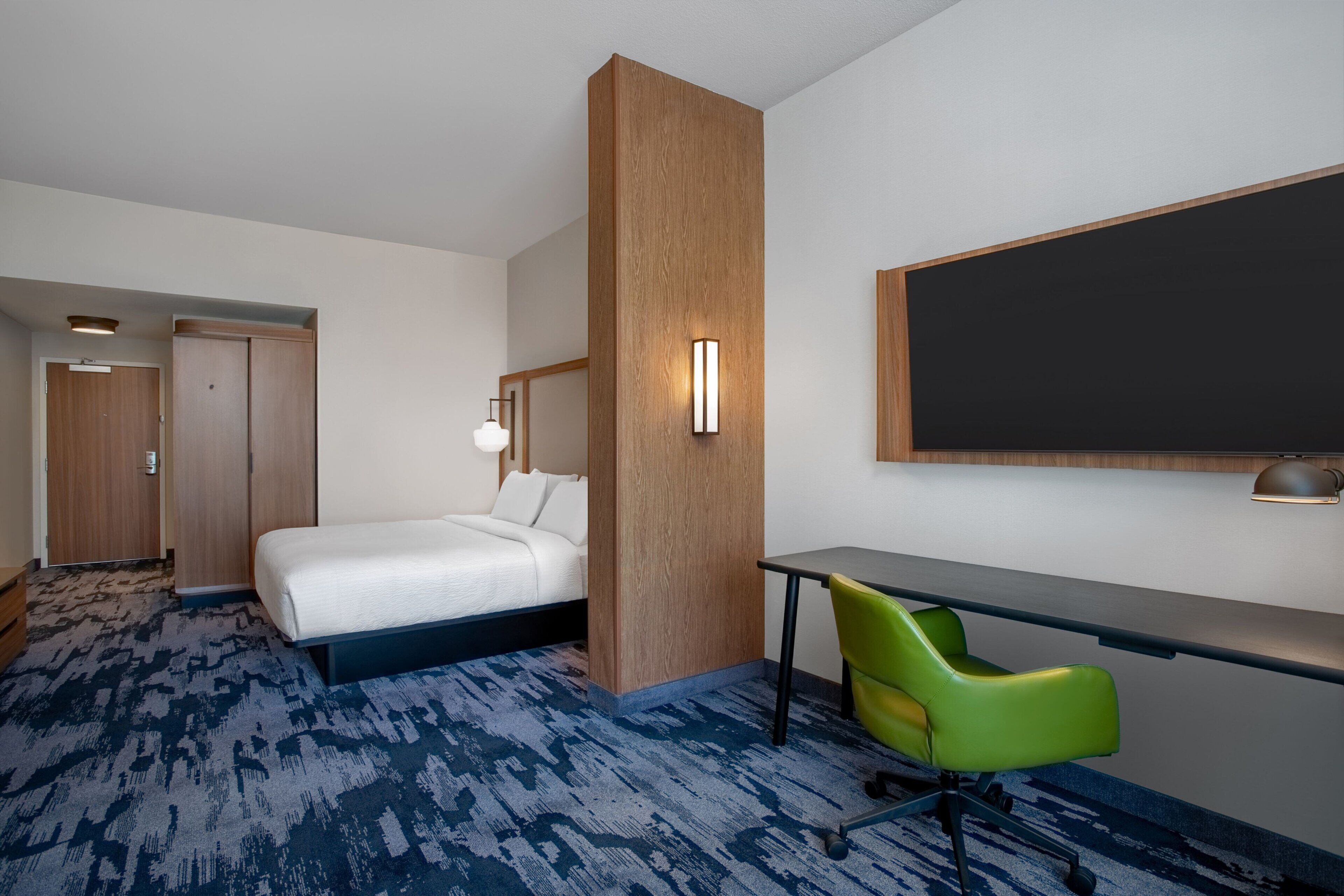 Habitació d'hotel amb un llit, una taula de treball, una cadira verda, una paret decorativa de fusta i un mirall gran.