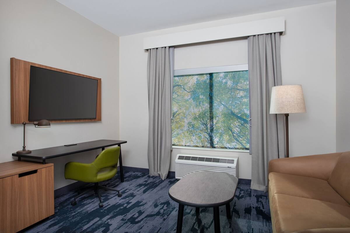 A hotel room with a desk, chair, sofa, TV, window with curtains, and a lamp.