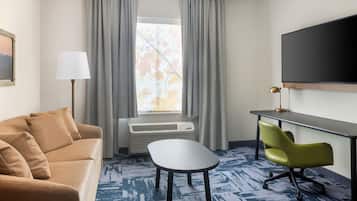 Ein Hotelzimmer mit einer Couch, einem kleinen Tisch, einem Schreibtisch mit Stuhl, einem Flachbildfernseher und einem Fenster mit Vorhängen.