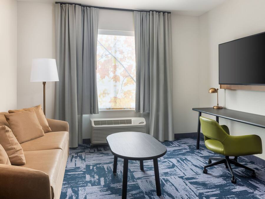 Ein Hotelzimmer mit einer Couch, einem kleinen Tisch, einem Schreibtisch mit Stuhl, einem Flachbildfernseher und einem Fenster mit Vorhängen.