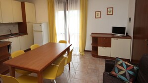Ein kompakter Wohnbereich mit Küchenzeile, einem Essbereich mit gelben Stühlen, einer Couch mit gemustertem Kissen und einem Fernseher auf einem Holzschrank.