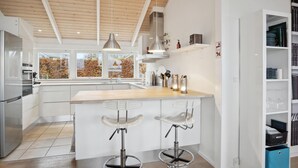 A modern kitchen with a central island, bar stools, pendant lighting, and a view of autumn foliage through large windows.