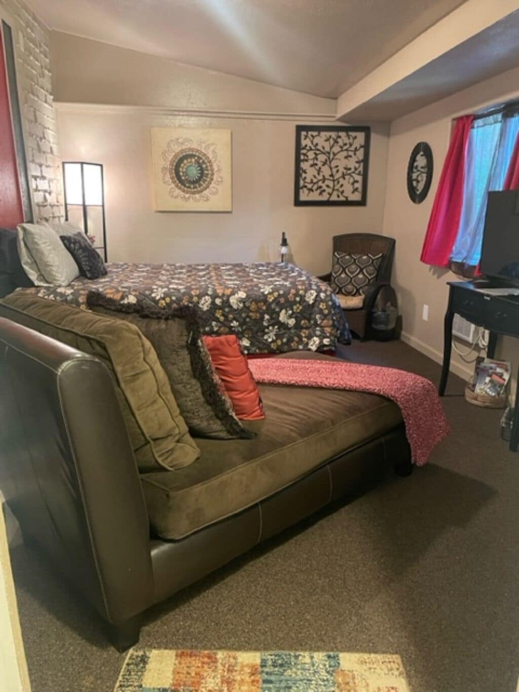 A cozy room with a large sofa, a small table, and a chair. There is a window with curtains, a wall clock, and a framed picture.