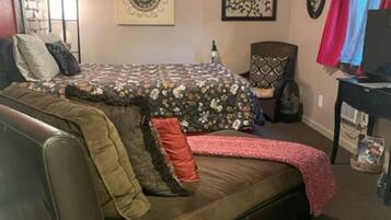 A cozy room with a large sofa, a small table, and a chair. There is a window with curtains, a wall clock, and a framed picture.