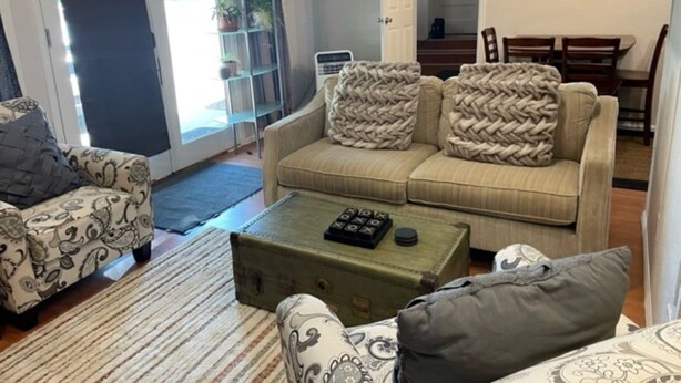 Un salon comprenant un canapé, un fauteuil et une table basse. Une porte donne accès à un balcon agrémenté de plantes.