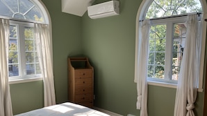 Ein Schlafzimmer mit zwei Fenstern, einem Bett, einer Holzkommode und einer Klimaanlage.