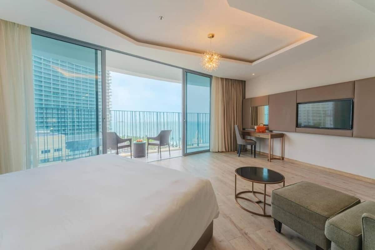 A modern hotel room with a large bed, a desk, a chair, and a TV. The room has large windows and a balcony with a view of the city.