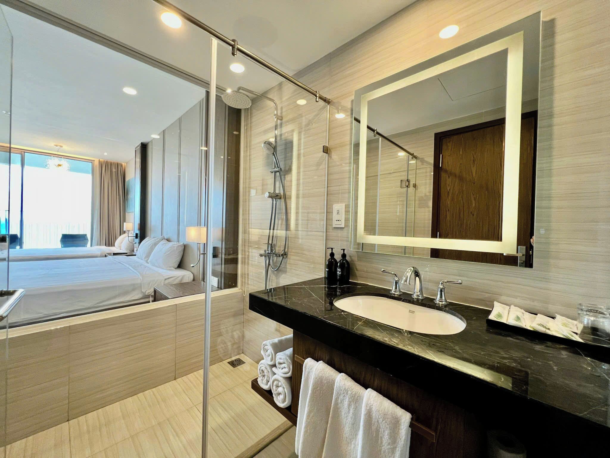 A modern hotel bathroom with a glass shower enclosure, a large mirror, a black marble sink, and a visible bed with a view to a balcony.