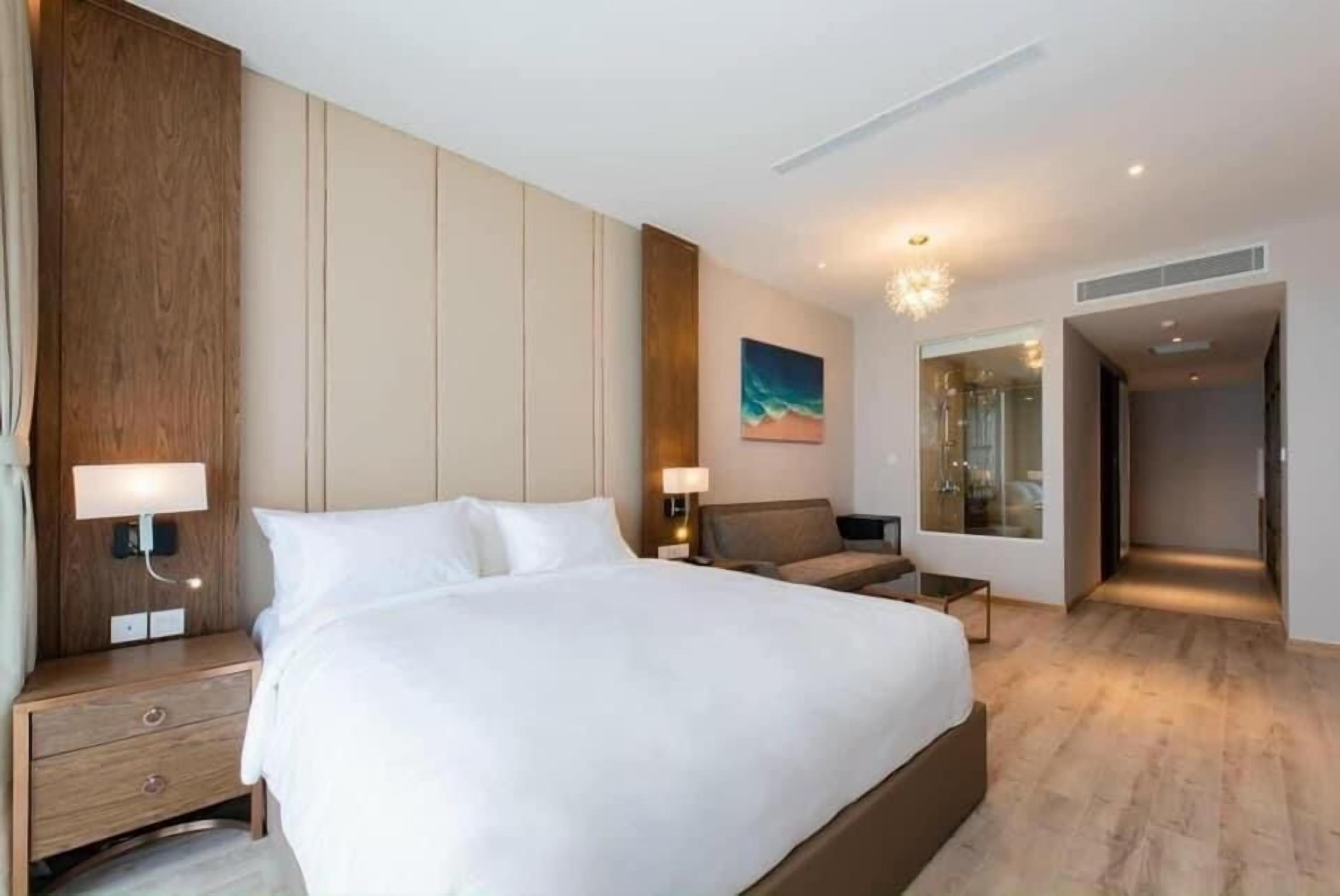 A modern hotel room with a large bed, bedside tables, a nightstand, a wall-mounted lamp, a painting, and a glass shower enclosure.