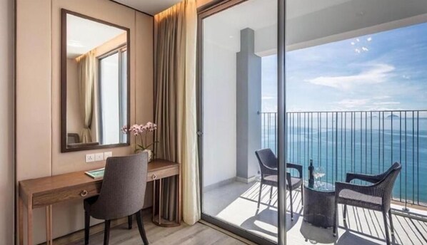 A hotel room with a desk, chair, and a view of the ocean through a balcony.