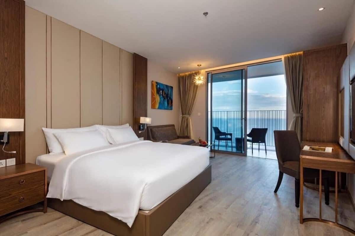 A modern hotel room with a large bed, a desk, and a balcony with a view.
