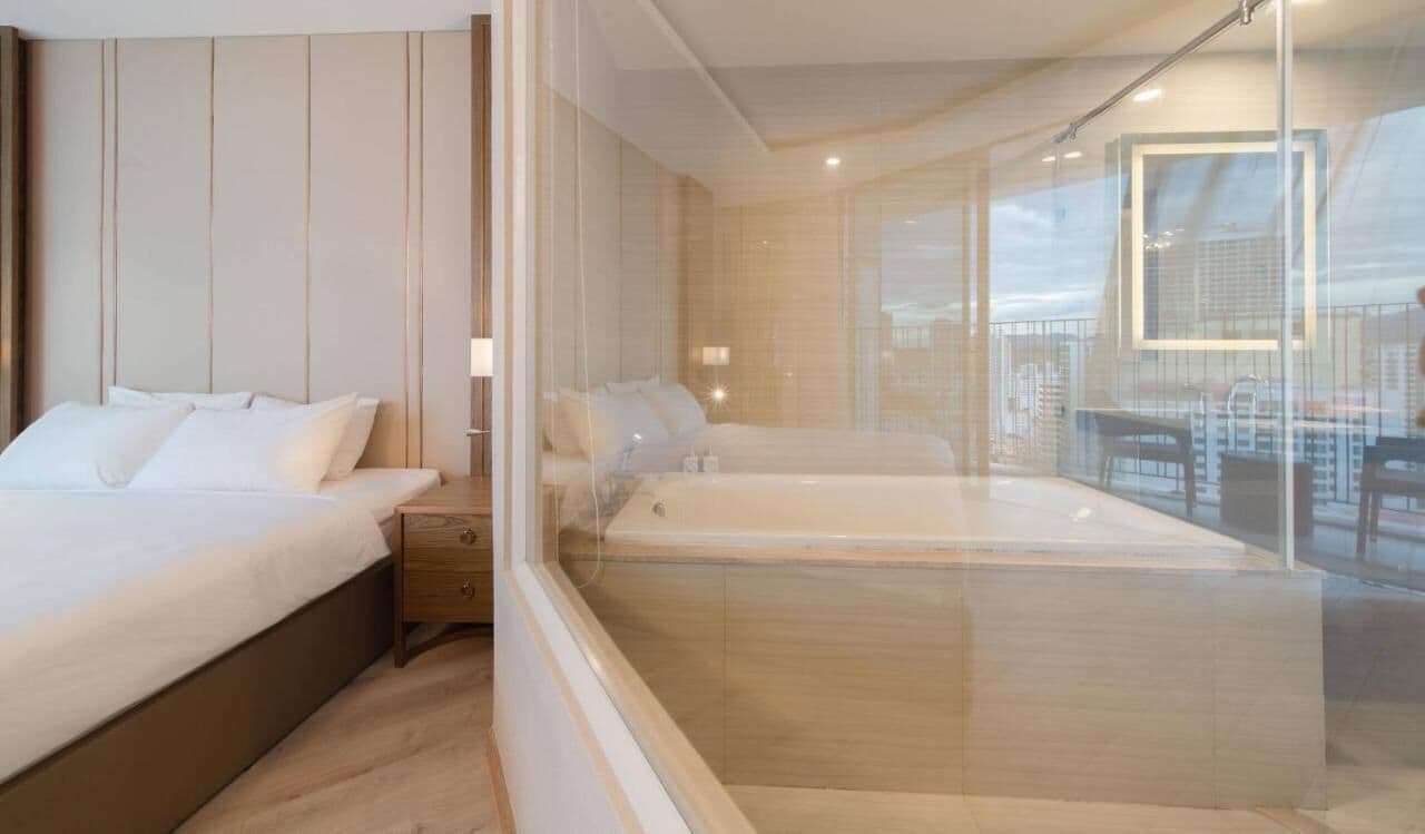 Quarto moderno com uma cama grande, área de estar separada e uma banheira de hidromassagem com vidro.
