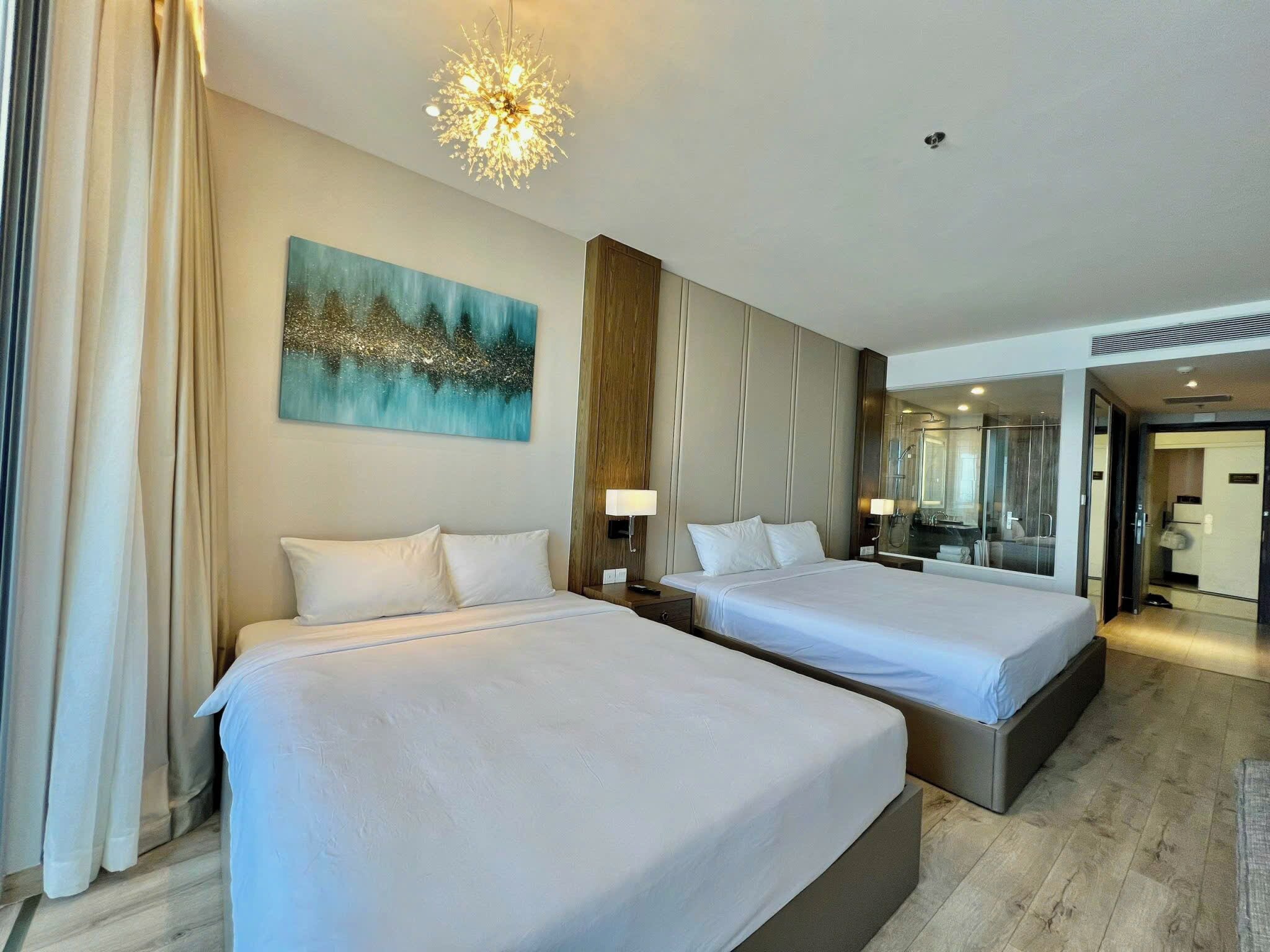 A hotel room with two beds, a large painting, a chandelier, and a bathroom visible through a glass door.