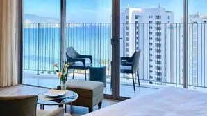 A hotel room with a large bed, two chairs, a small table, and a view of the ocean and buildings through floor-to-ceiling windows.