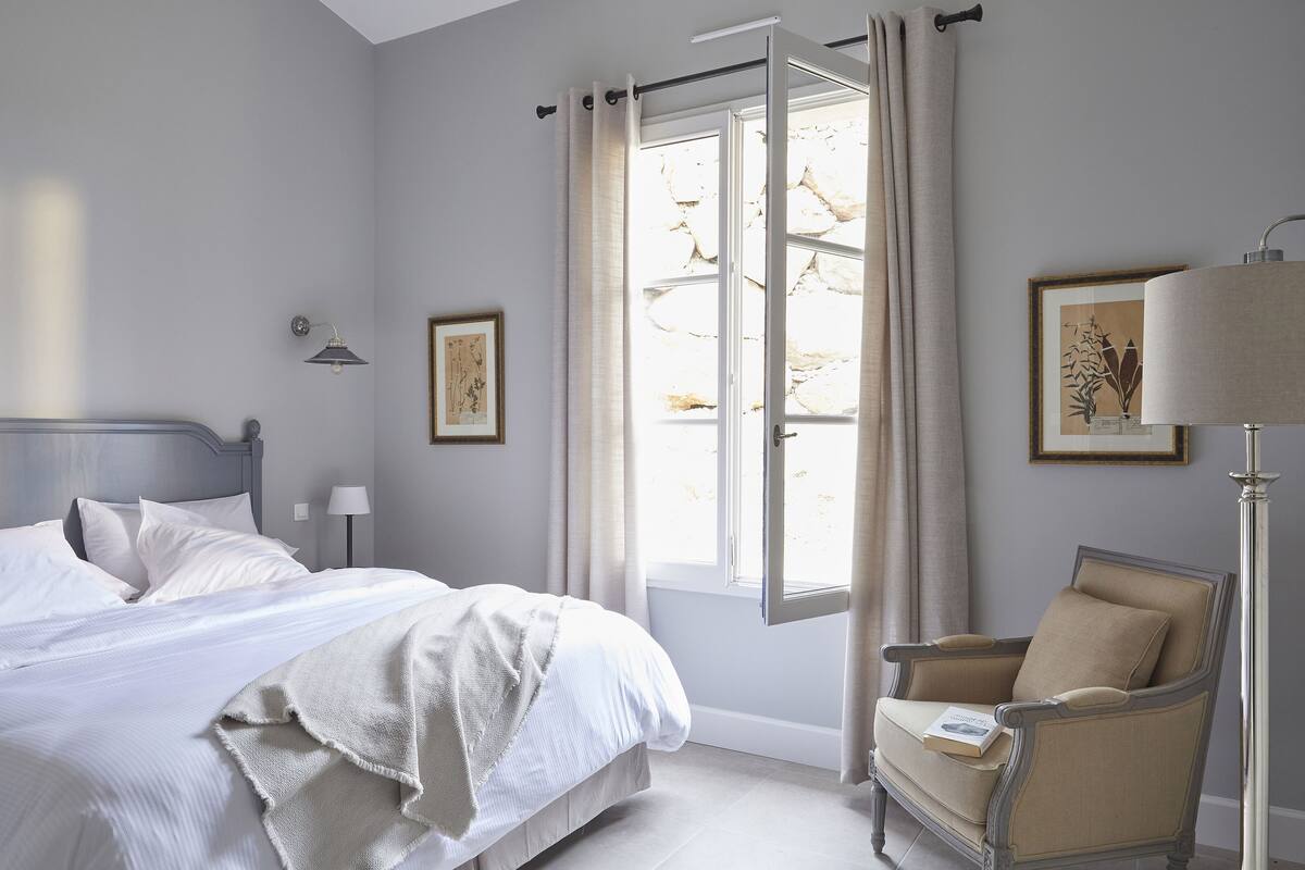Ein Schlafzimmer mit Bett, Stuhl, Lampe und einem Fenster mit Vorhängen.