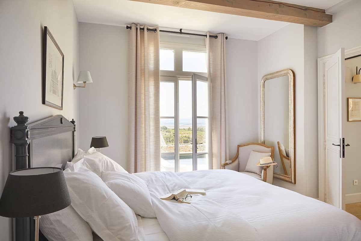 Ein Schlafzimmer mit einem Bett, einem Nachttisch, einer Lampe, einem Spiegel und einem Fenster mit Vorhängen.