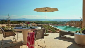 Um pátio com mesa redonda, cadeiras de vime, guarda-sol e vista para uma piscina e paisagem.