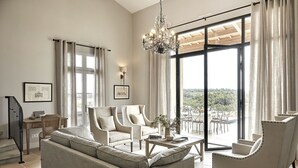 Ampla sala de estar com um sofá grande, duas poltronas, uma mesa de centro e um lustre. Há uma escada com corrimão, uma área de jantar com cadeiras e uma janela grande com cortinas.