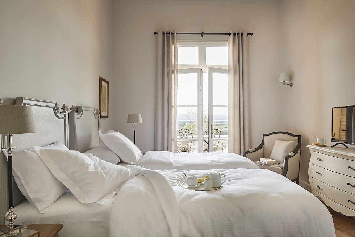 Ein Hotelzimmer mit einem großen Bett, einem Nachttisch mit Lampe, einem Schminktisch, einem Sessel und einem Fenster mit Vorhängen.