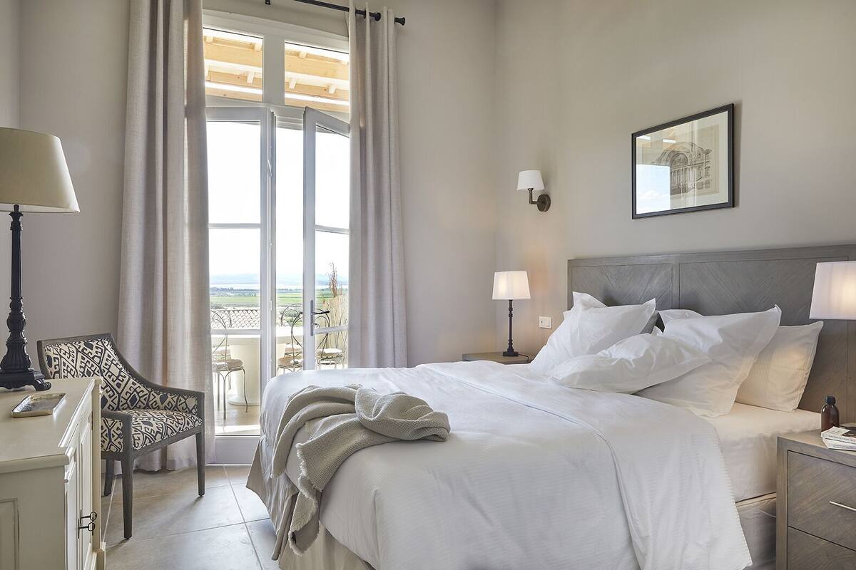 Ein Hotelzimmer mit Bett, zwei Nachttischen, einem Sessel und Schreibtisch sowie Blick ins Freie.