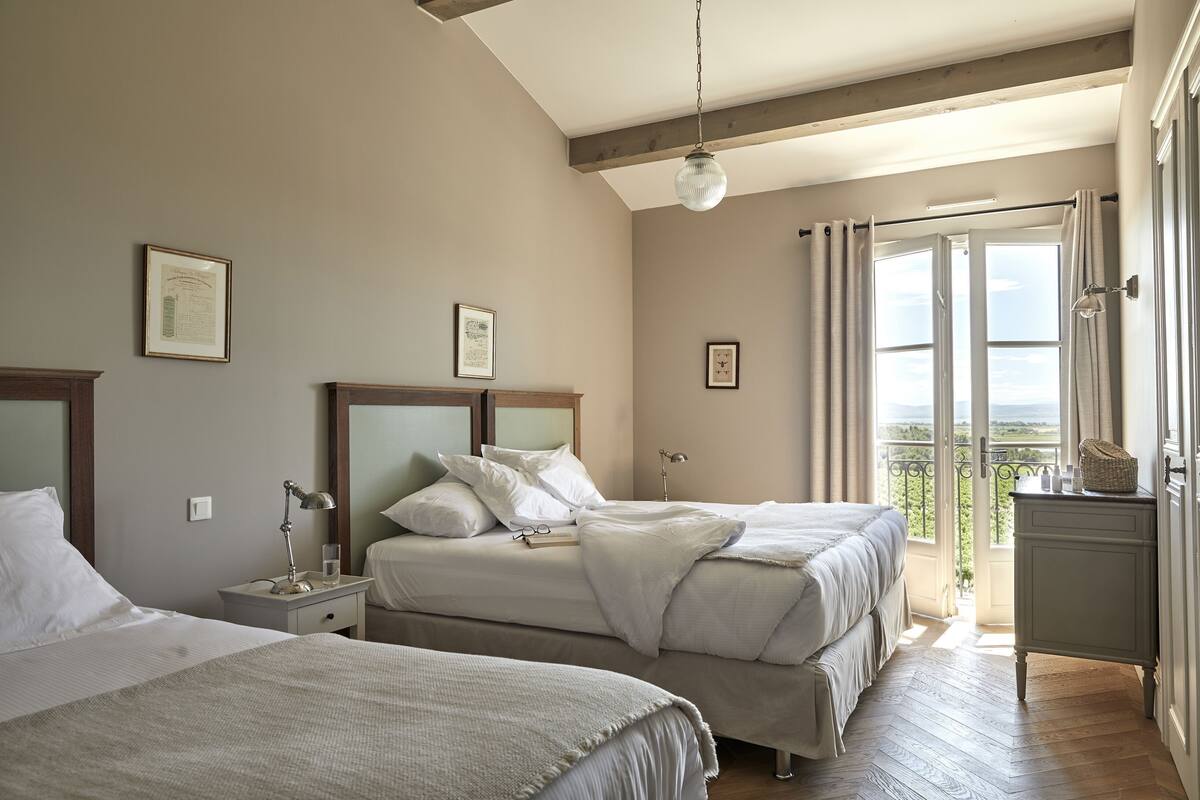Ein Schlafzimmer mit zwei Betten, einem Nachttisch, einer Lampe und Blick ins Freie.