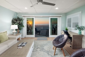 Un salon moderne avec un canapé, une table basse et un fauteuil. On y trouve un bureau avec un ordinateur et, grâce à des portes vitrées, une vue sur la plage.