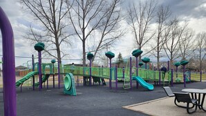 Une aire de jeux dotée d’équipements verts et violets, notamment des toboggans et des structures d’escalade, et entourée d’arbres.