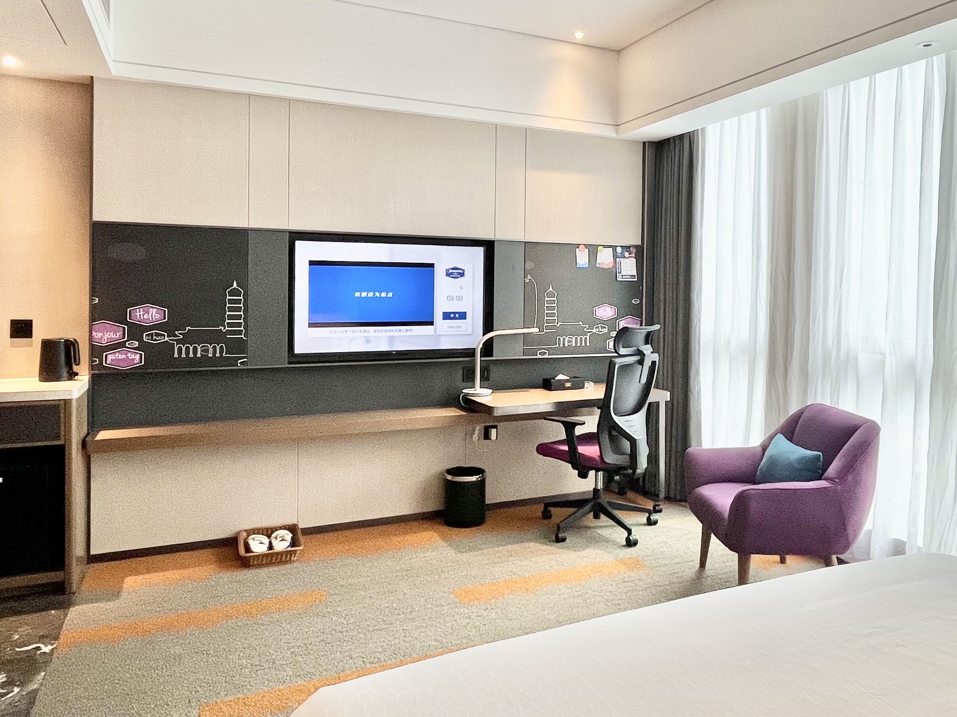 A hotel room with a bed, a desk with a computer, a chair, a TV mounted on the wall, and a window with curtains.