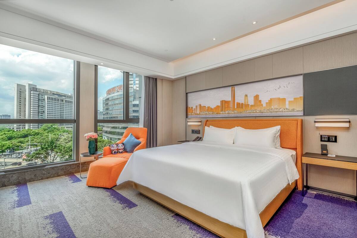Una habitación de hotel moderna con una cama grande, vistas a la ciudad, un sillón cómodo y un escritorio con lámpara.