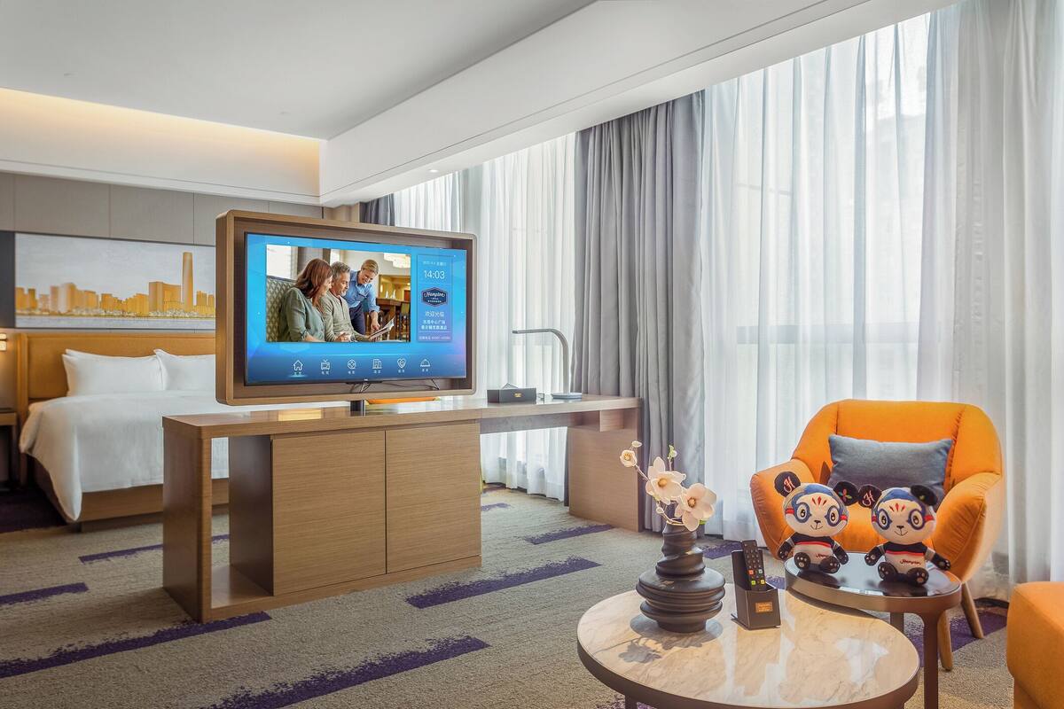 Habitación de hotel con un televisor mostrando un noticiero, un sofá, un sillón naranja, una mesita con un florero y una cama con ropa de cama blanca.