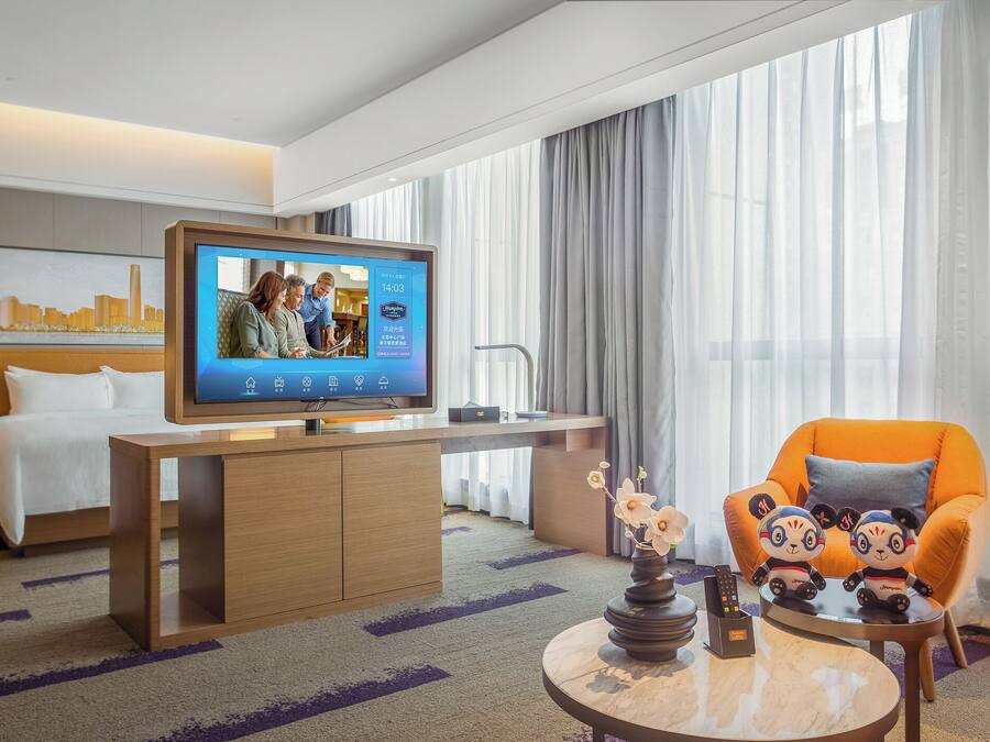 Habitación de hotel con un televisor mostrando un noticiero, un sofá, un sillón naranja, una mesita con un florero y una cama con ropa de cama blanca.