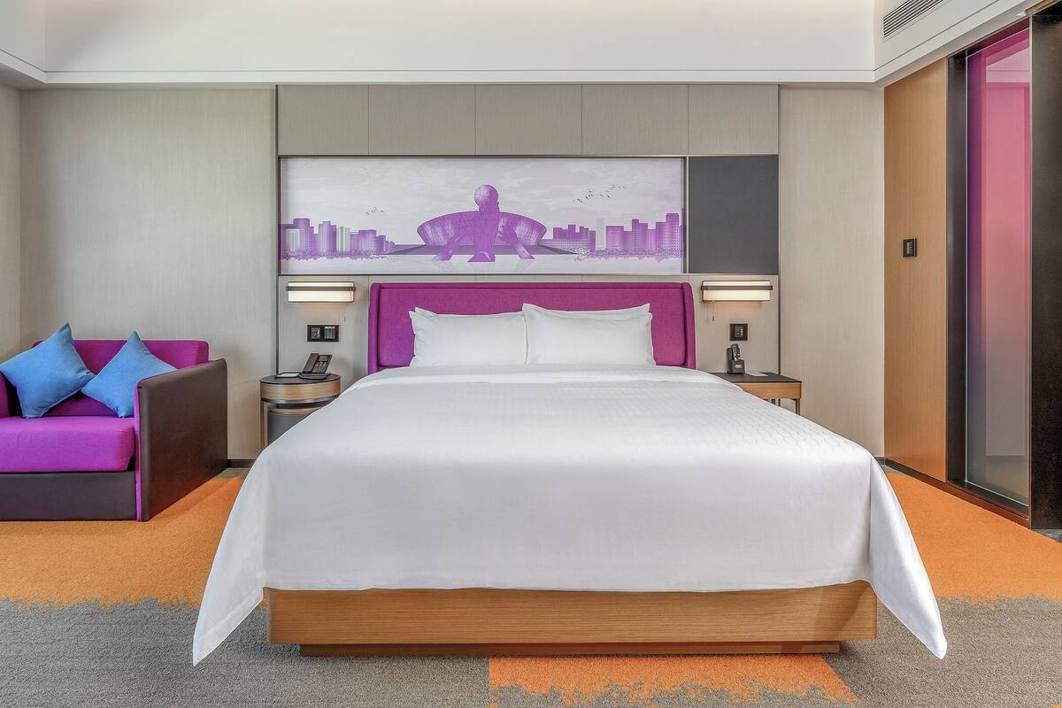 Una habitación de hotel moderna con una cama grande, una obra de arte urbana sobre la cama, un cabecero morado, una mesita de noche con un teléfono y un sofá rosa con almohadas azules.