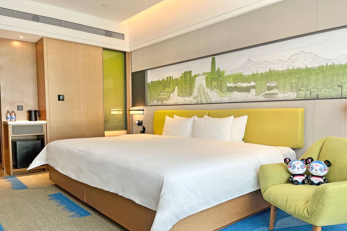 Una habitación de hotel con una cama grande, un sillón verde, un mural con vista a la ciudad y un mini bar.