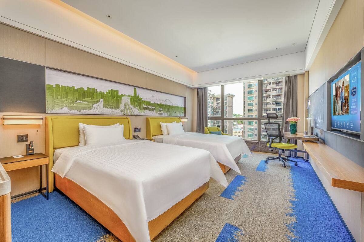 Habitación de hotel con dos camas, un escritorio, televisor y un ventanal con vistas a la ciudad.