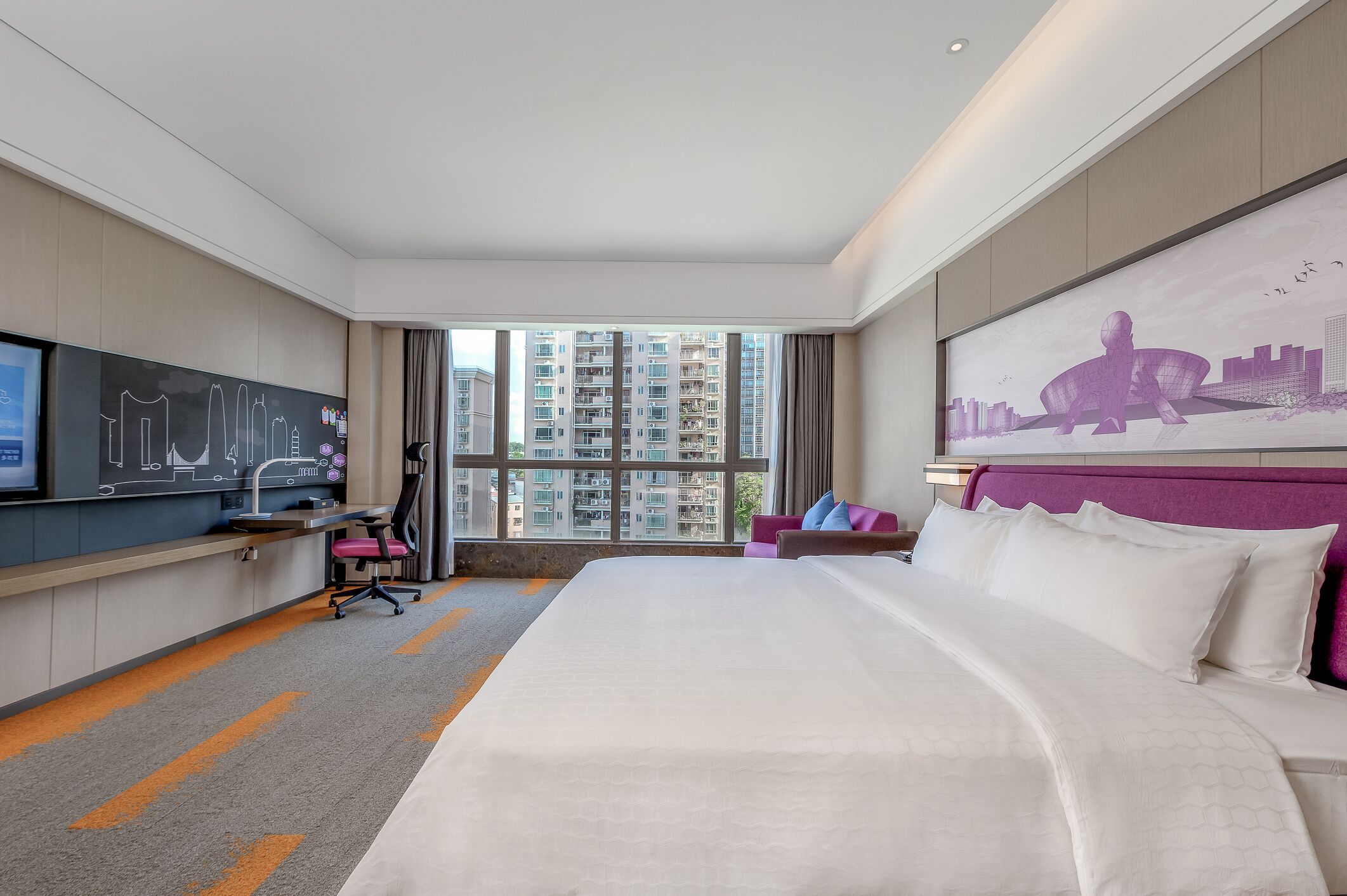 A hotel room with a large bed, a desk with a computer, a chair, and a window with a city view.