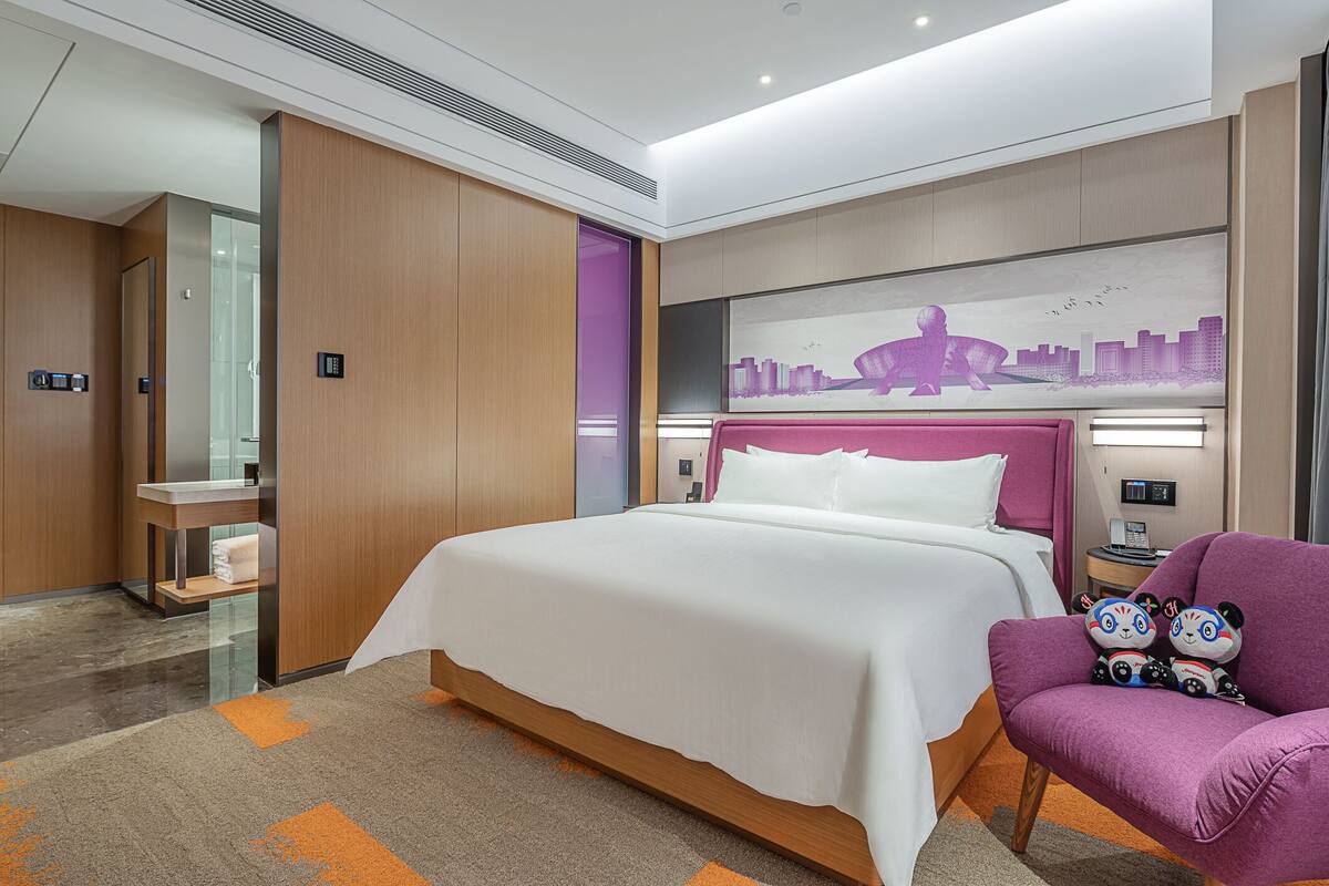 Una habitación de hotel moderna con una cama grande, un tocador con espejo, un sillón púrpura y un mural con vista a la ciudad.