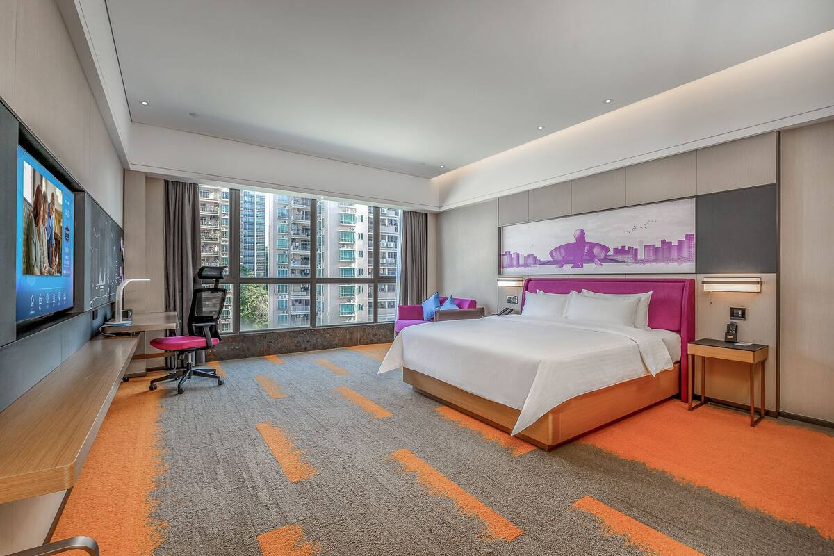 Una habitación de hotel moderna con una cama grande, un escritorio con una silla, un televisor y un cuadro de paisaje urbano en la pared.