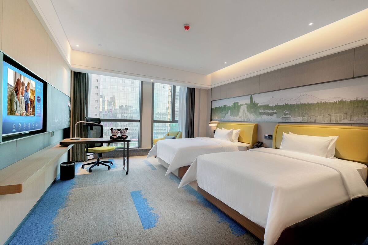 Habitación de hotel con dos camas, un escritorio con computadora, televisión y un ventanal con vista a la ciudad.