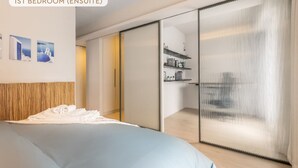 Une chambre moderne, avec un grand lit, dotée d’une porte-fenêtre coulissante donnant sur une salle de bain, d’une tête de lit en bois et d’un lit agrémenté de linge de lit blanc.