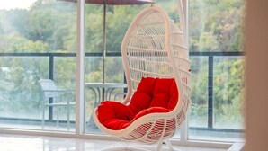 Una silla mecedora de mimbre blanco con cojines rojos en un balcón con barandilla de vidrio, con vistas a un jardín.