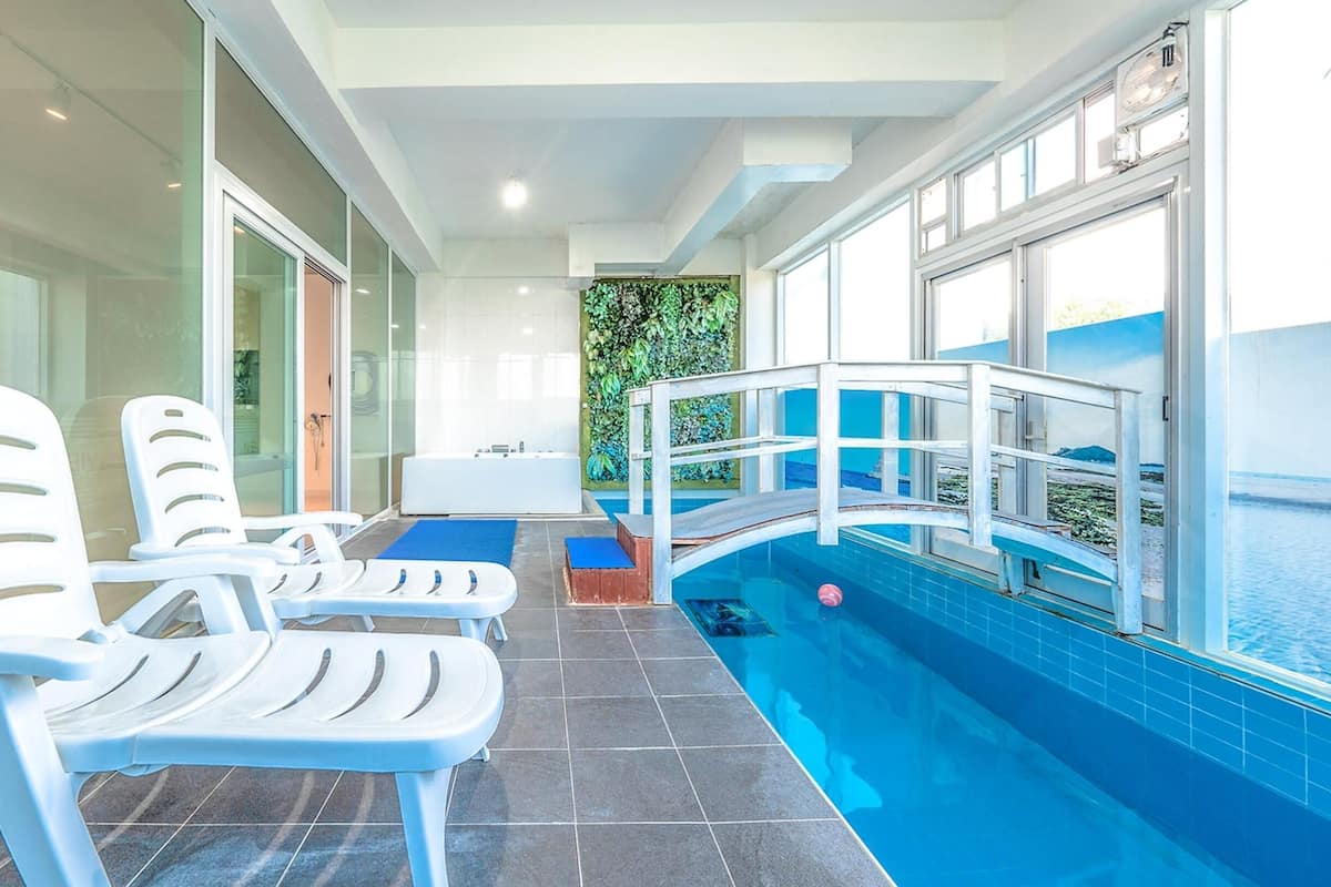 A swimming pool area with a diving board, lounge chairs, and a glass-enclosed room with a view of the sea.