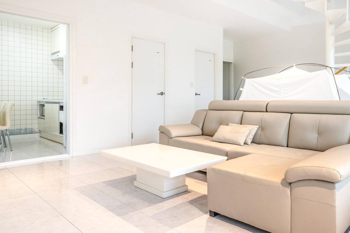 A modern living room with a beige sofa, a white coffee table, and a glass door leading to a kitchen.